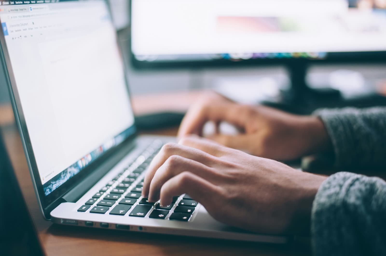 Hände tippen auf der Tastatur eines Laptops, dahinter ein zweiter Bildschirm