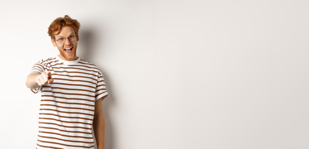 Image of funny redhead guy in glasses laughing at you, pointing finger camera and making fun of someone, standing over white background