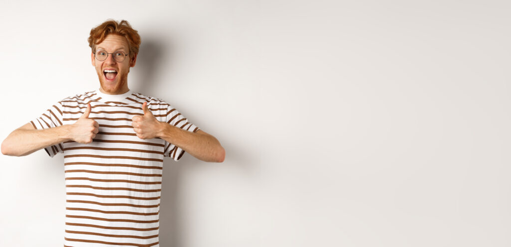 Young amazed man in red hair checking out something awesome, saying yes and showing thumbs-up, standing over white background