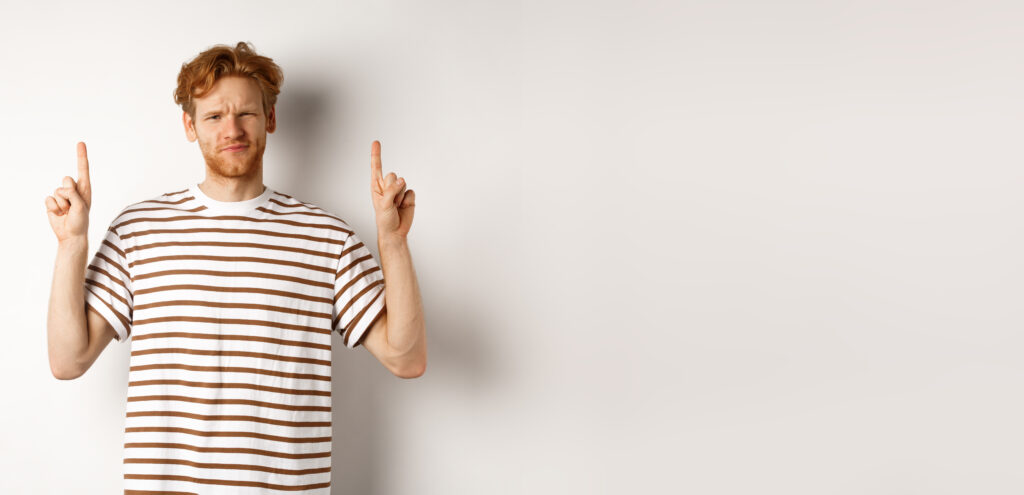 Skeptical handsome redhead man pointing fingers up, grimacing and looking doubtful at camera, standing indecisive over white background