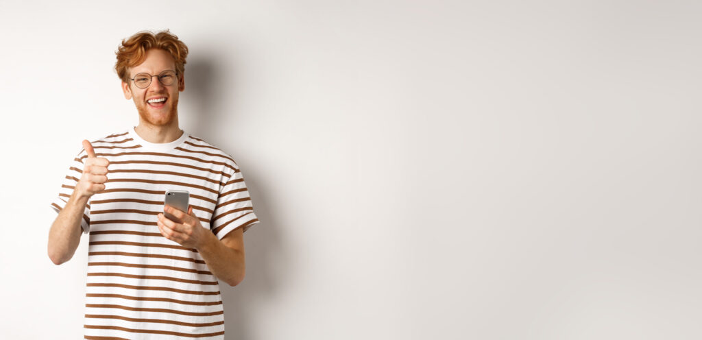 Technology and e-commerce concept. Satisfied male model with red hair, showing thumbs-up and holding smartphone, white background
